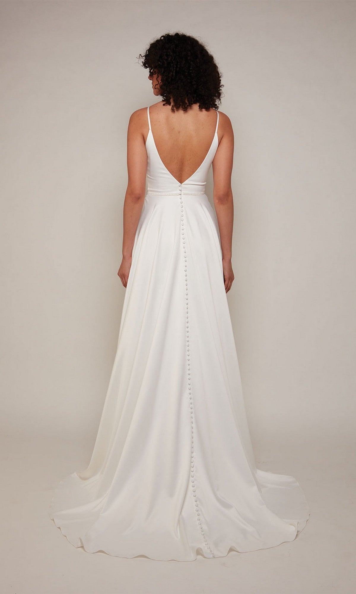 A woman with curly hair models the Alyce 7089 long formal dress, a minimalist A-line bridal gown with a deep V-back and button detailing, posed against a plain background.